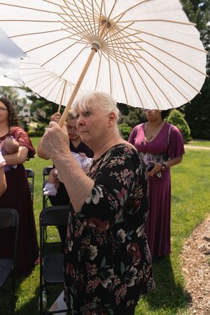 Elderly woman in floral dress holding white paper parasol at outdoor wedding ceremony.