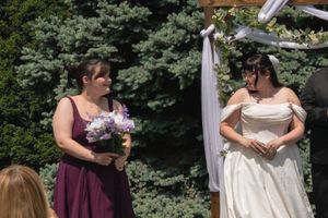 Bridesmaid in purple dress holding bouquet stands beside bride in white gown at outdoor wedding altar.
