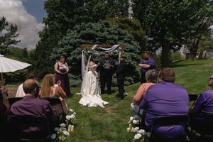 Outdoor garden wedding ceremony with bride and groom standing under floral arch with guests.