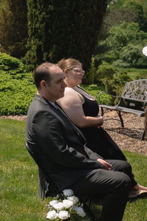 Man in gray suit and woman in black dress seated as guests at outdoor wedding.