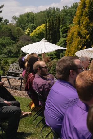 Outdoor wedding ceremony guests seated in chairs with white parasols and lush green garden backdrop on sunny day.
