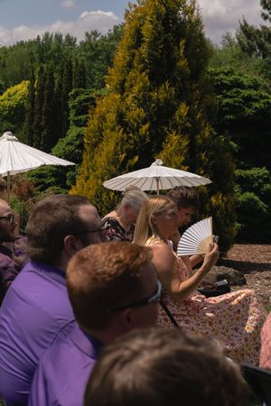 Wedding guests seated outdoors in summer heat, woman using hand fan near decorative parasol umbrellas.