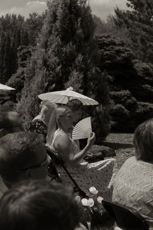 Black and white photo of a bride kissing her groom under a parasol at an outdoor wedding ceremony.