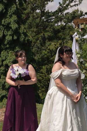 Bridesmaid in burgundy gown stands beside bride in white off-shoulder dress at outdoor ceremony.