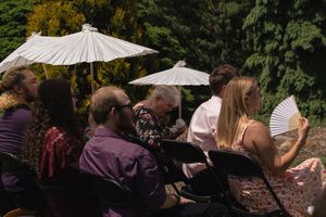 Wedding guests seated outdoors shade themselves with white parasols and hand fans during an outdoor ceremony.