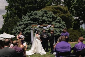 Outdoor wedding ceremony with bride in white gown at floral arch, guests seated on lawn surrounded by lush green trees.