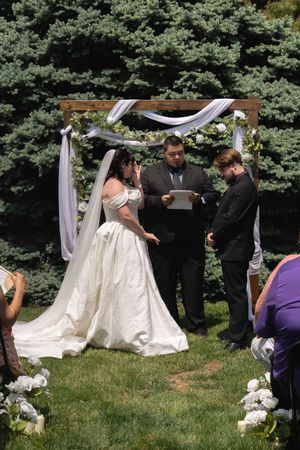Bride in white gown and groom exchange vows at outdoor wooden arch wedding ceremony altar.
