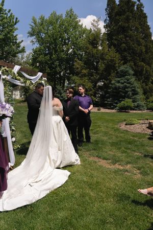 Bride in white gown and veil stands at outdoor wedding ceremony altar surrounded by guests on green lawn.