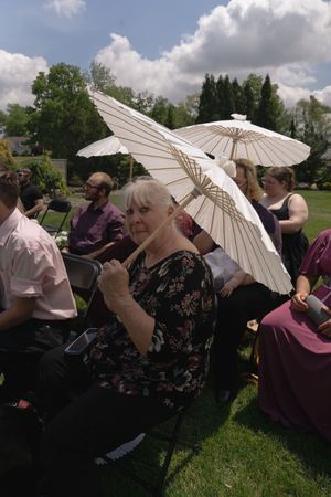 Outdoor wedding guests seated on lawn holding white paper parasol umbrellas on cloudy day.