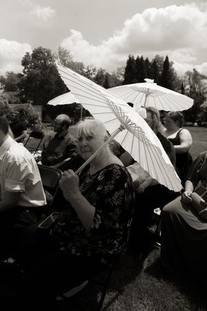 Black and white photo of wedding guests seated outdoors holding white parasol umbrellas during ceremony.