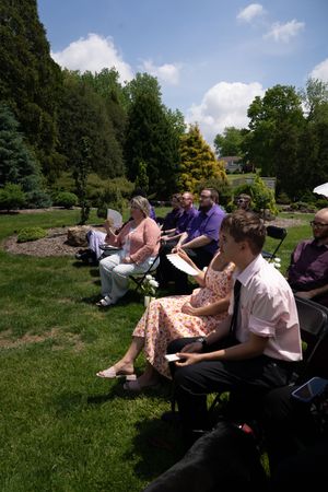 Wedding guests seated in chairs on sunny lawn fan themselves during outdoor garden ceremony.