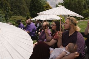 Wedding guests seated outdoors holding white parasols for shade during sunny garden ceremony.
