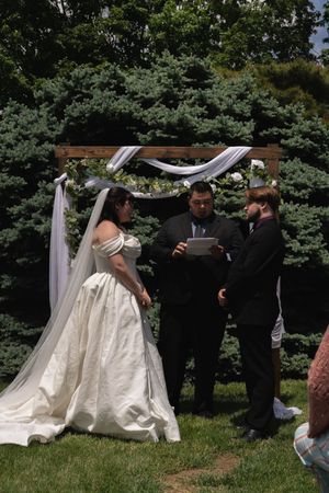 Bride and groom exchange vows under decorated wooden arch with white draping outdoors.
