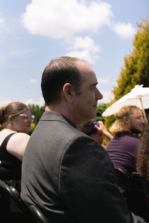 A man in a gray suit sits among wedding guests at an outdoor ceremony with white parasols in the background.