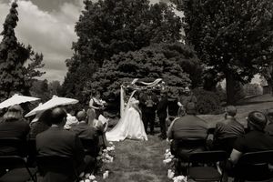 Black and white wide shot of outdoor wedding ceremony with seated guests and floral aisle decorations.