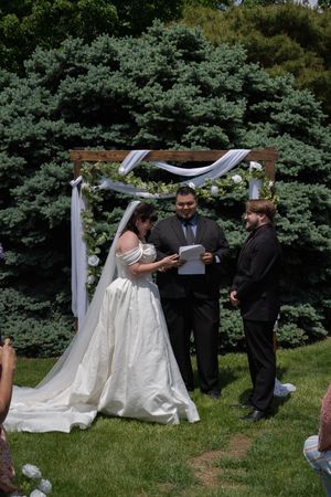 Bride and groom exchanging vows outdoors under a wooden arch with white draping and flowers, surrounded by lush green trees.