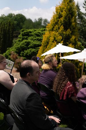 Guests seated outdoors at a wedding ceremony, with white parasols and lush green trees in the background.