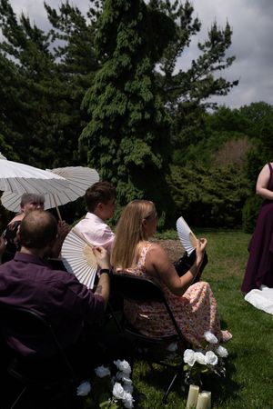 Wedding guests use white parasols and fans to stay cool during outdoor garden ceremony.