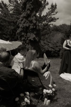 Black and white photo of wedding guests using hand fans and parasol during outdoor garden ceremony.