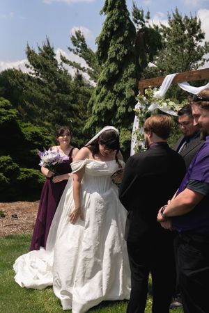 Bride in white gown bows head during outdoor wedding ceremony beside bridesmaid in purple dress near floral arch.