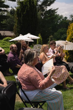 Wedding guests seated outdoors in sunny garden using parasols and fans to stay cool.