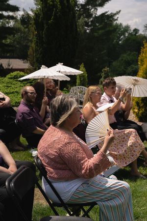 Wedding guests seated outdoors using parasols and fans to shade from summer sun.