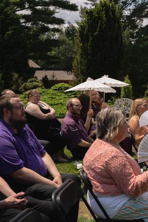 Wedding guests seated outdoors in garden ceremony, wearing purple attire with parasol visible.