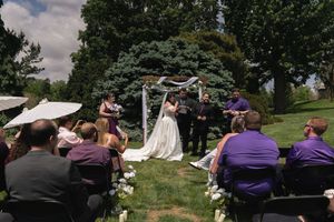 A bride and groom share a kiss at an outdoor wedding ceremony as guests in purple attire look on joyfully.