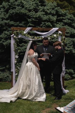 Officiant reads vows as bride and groom stand under white-draped wooden wedding arch outdoors.