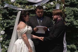 Bride and groom exchange vows holding hands beneath floral arch with officiant reading ceremony script.