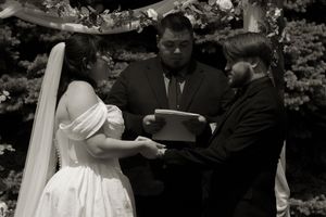Black and white photo of bride and groom holding hands during outdoor wedding ceremony with officiant.