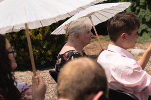 Wedding guests use white parasols for shade during outdoor ceremony on sunny day.