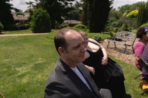 Man in gray suit seated at outdoor wedding ceremony in sunny garden setting.