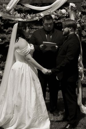 Black and white photo of couple holding hands at altar as officiant reads vows under decorated wedding arch.