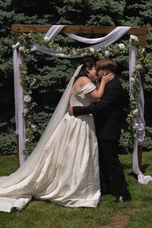 Bride and groom sharing first kiss under rustic wooden arch adorned with white roses and draping.