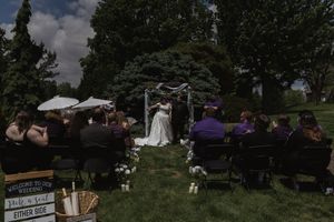 Outdoor wedding ceremony with guests seated on chairs facing bride and groom at floral arch.