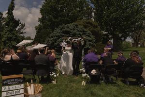 Outdoor wedding ceremony on green lawn with guests seated in black chairs facing couple under floral arch.