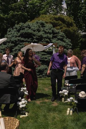 Bridesmaid in burgundy dress and groomsman walk down outdoor wedding aisle after ceremony.