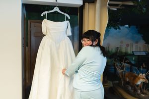 Woman in light blue outfit carefully adjusts a white wedding dress hanging on a door hanger indoors.