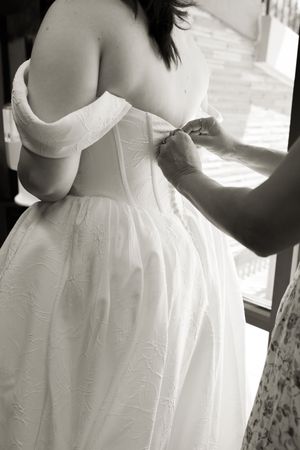 Black and white close-up of bride having wedding dress buttoned up before ceremony.