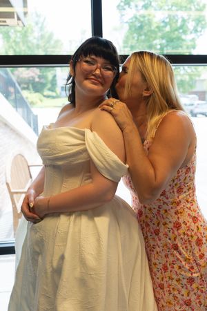 Blonde woman in floral dress kisses bride on cheek indoors before wedding ceremony.