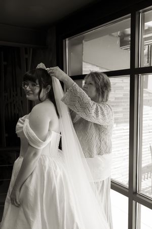 Black and white photo of woman helping bride secure veil before wedding ceremony indoors.