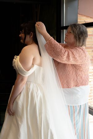 Older woman in pink sweater helping bride put on wedding veil before ceremony.