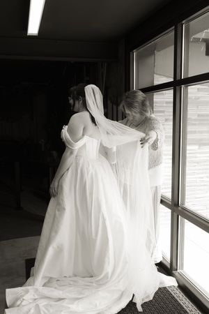 Black and white photo of bride in off-shoulder gown being helped with veil near window before wedding ceremony.