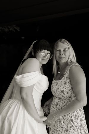 Black and white portrait of bride in white gown and veil posing with blonde woman indoors.