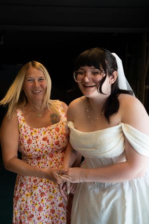 Laughing bride in white off-shoulder gown with veil posing alongside smiling woman in floral dress.