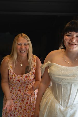 A bride in a white strapless gown laughs joyfully with a blonde woman in a floral dress at a wedding.