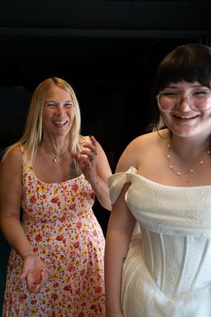 Joyful bride in white dress laughs with smiling blonde woman in floral dress at wedding reception.