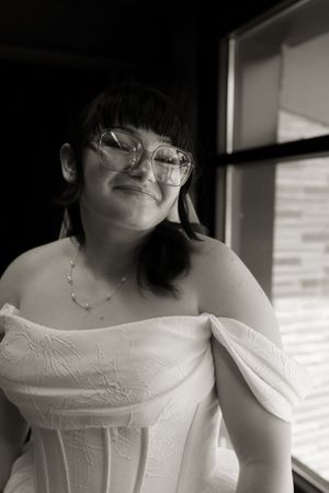 Black and white portrait of smiling bride in off-shoulder gown wearing glasses near window.