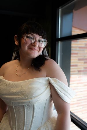 Smiling bride in an off-shoulder white corset wedding gown and glasses posing near a window indoors.